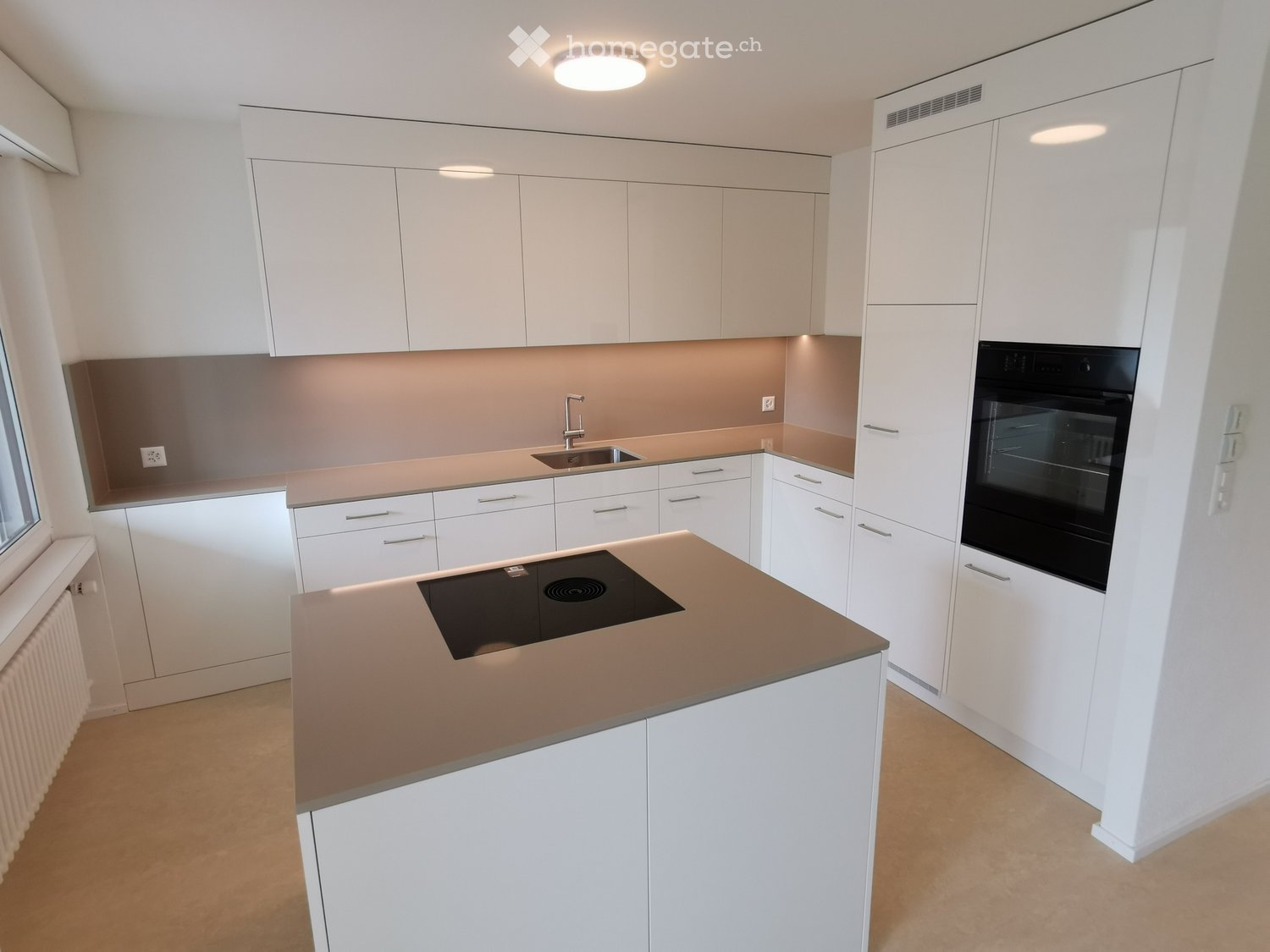 Modern, open-concept kitchen with white cabinets, beige countertops, built-in appliances including a dishwasher, and a central island with a cooktop.