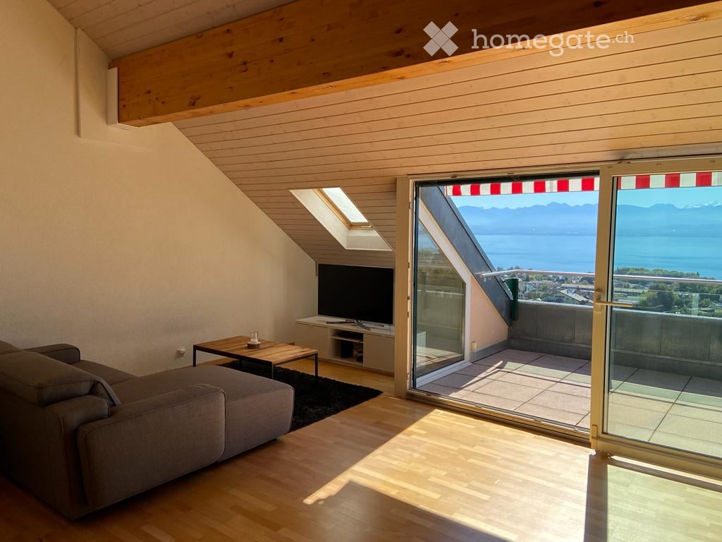 Modern living room with wooden floors, large windows, and a view of a lake and mountains. Features a grey sofa, coffee table, and TV.
