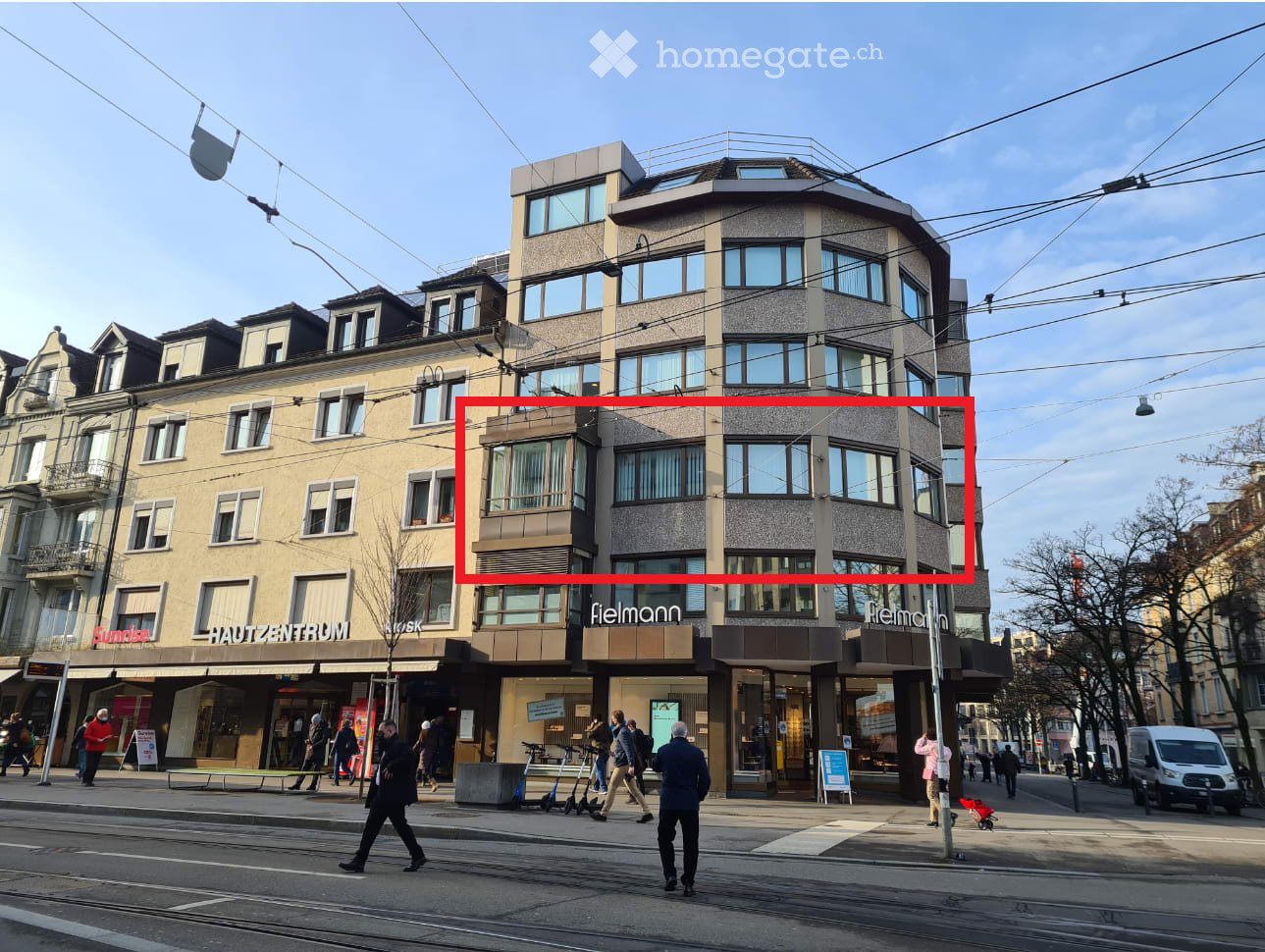 Corner building with several floors, gray exterior, large windows, stores on the ground floor
