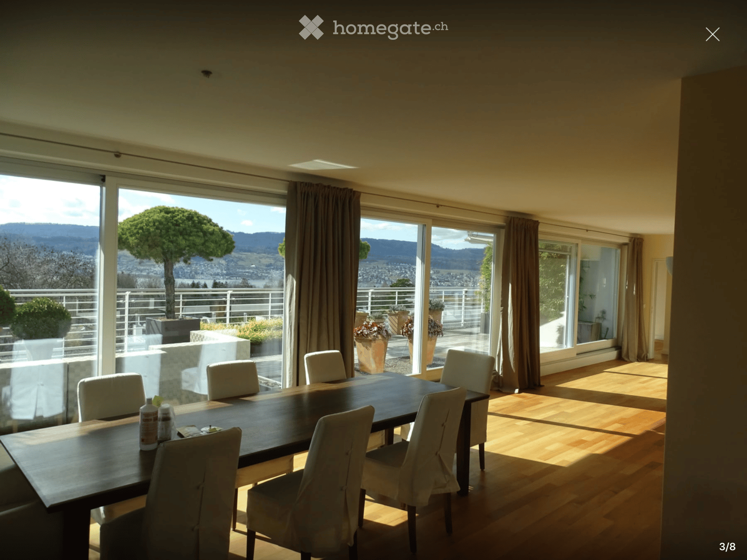 Dining room with a large wooden table and white chairs, large windows offering a scenic view of mountains and greenery, and a balcony area with potted plants visible through the windows.