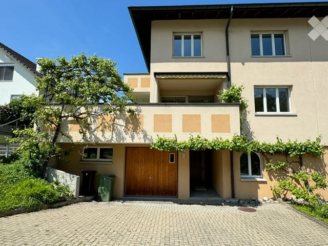 Modern two-story house with beige facade, large windows, wooden garage door, and a small balcony on the upper floor.