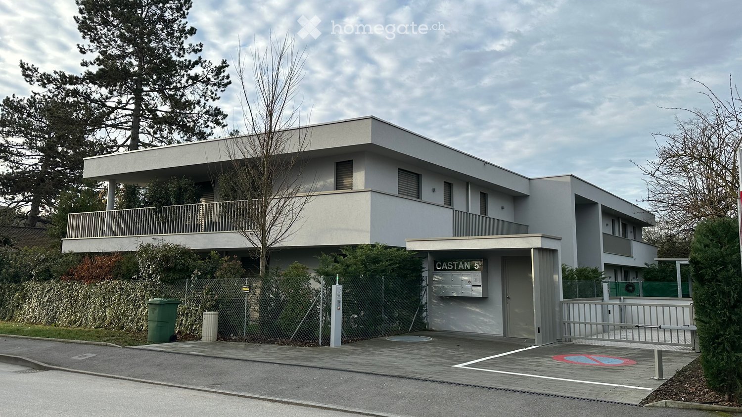 Modern two-story building with a flat roof, balconies, and a gated entrance area. The building has a clean, minimalist design with large windows and a small garden area in front.