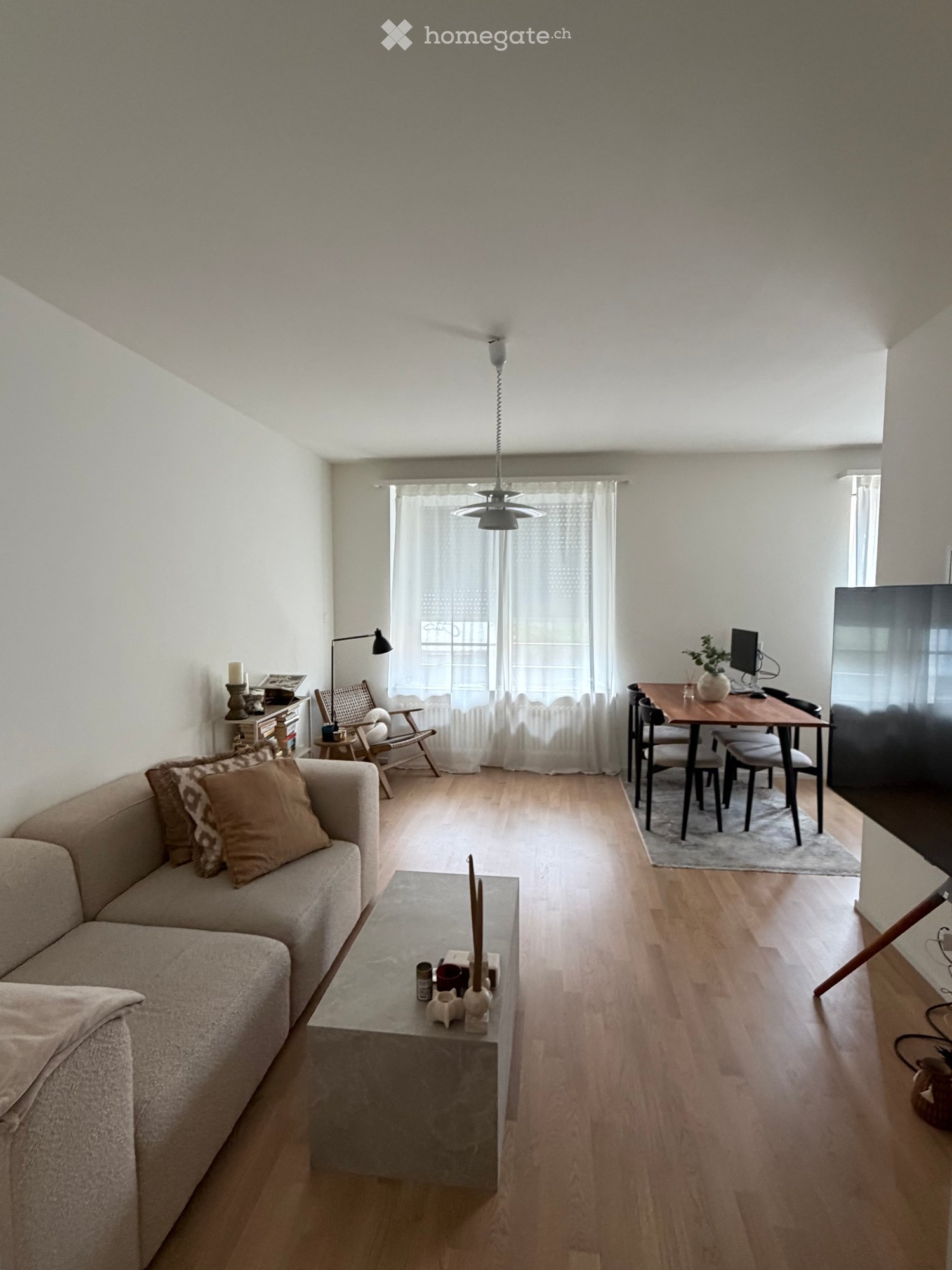 A modern living room with a sofa, coffee table, dining area, and a TV screen. The room features wooden flooring, large windows with sheer curtains, and a cozy seating area.