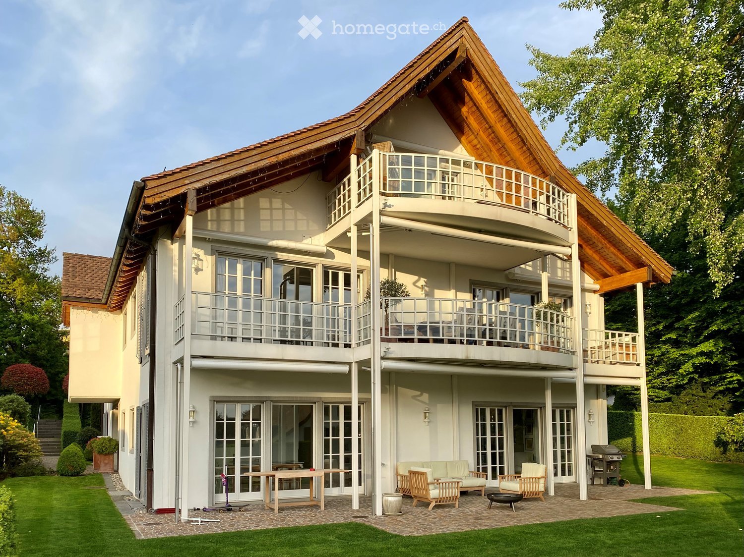 Two-story house with a wooden roof, balconies, and a well-maintained garden with outdoor seating and a grill.