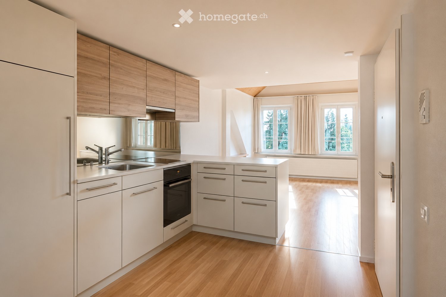 Modern kitchen with white cabinets, wooden accents, stainless steel sink, and built-in oven. The space features light wooden flooring and large windows with curtains.