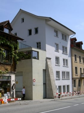 Modern multi-story building with white facade, large windows, and a small balcony on the second floor. The building has a contemporary design with clean lines and a mix of materials including concrete and glass.