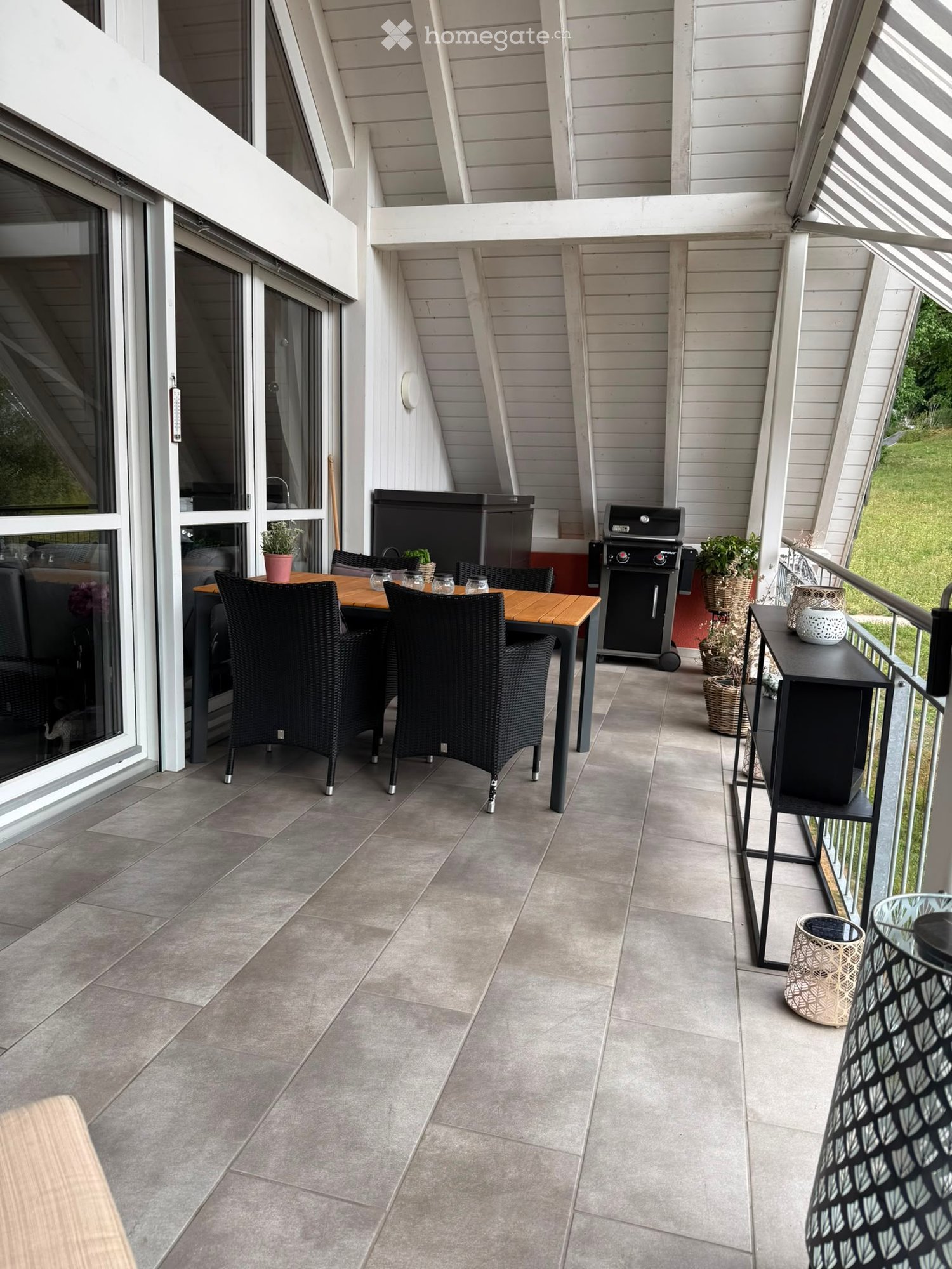 Balcony with tiled floor, wooden table, black wicker chairs, grill, potted plants, and side table.
