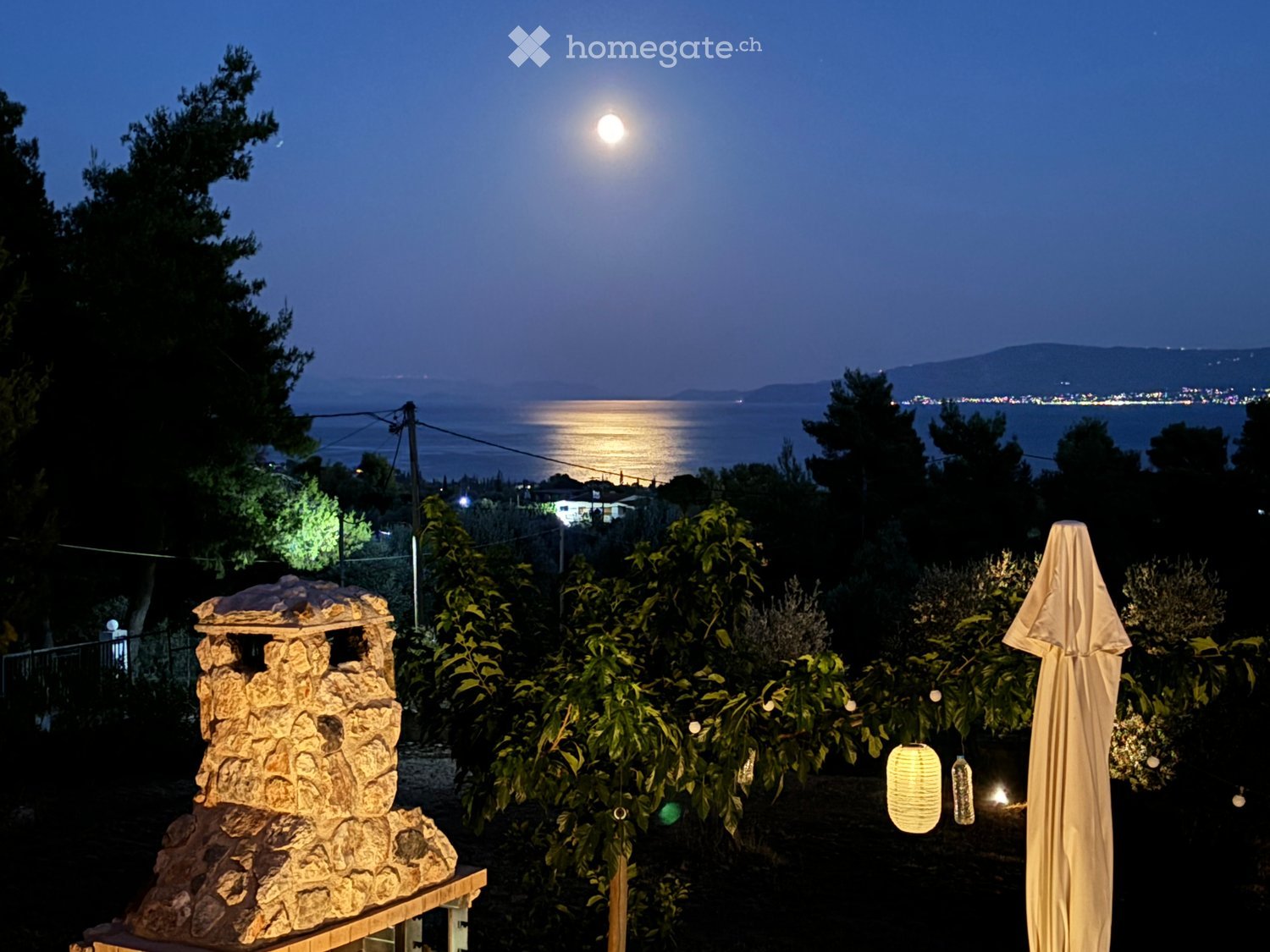 Nighttime garden with a stone structure, covered umbrella, string lights, and a view of the sea and distant city lights.