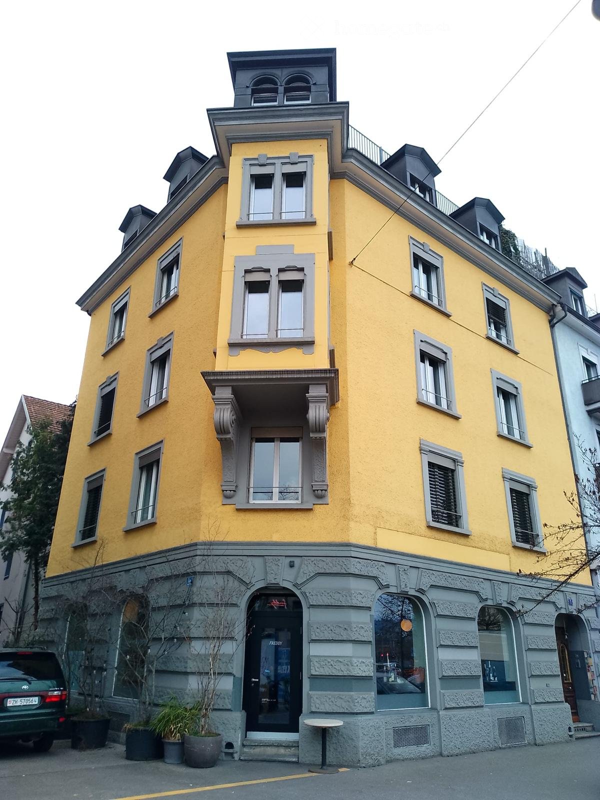 A multi-story yellow building with grey accents, featuring multiple windows, a central entrance with a small balcony, and a storefront with glass windows and a door labeled "FREEDY".