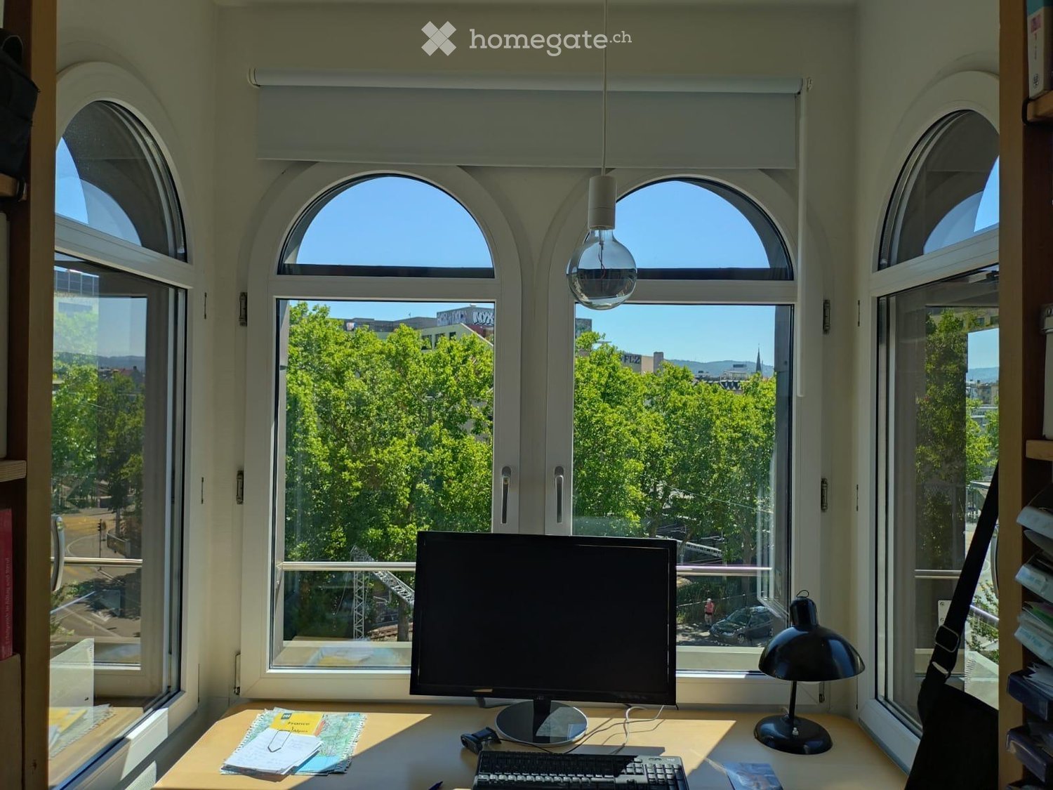 Brightly lit room with large arched windows, a wooden desk with a computer setup, a black desk lamp, and a view of greenery and cityscape.