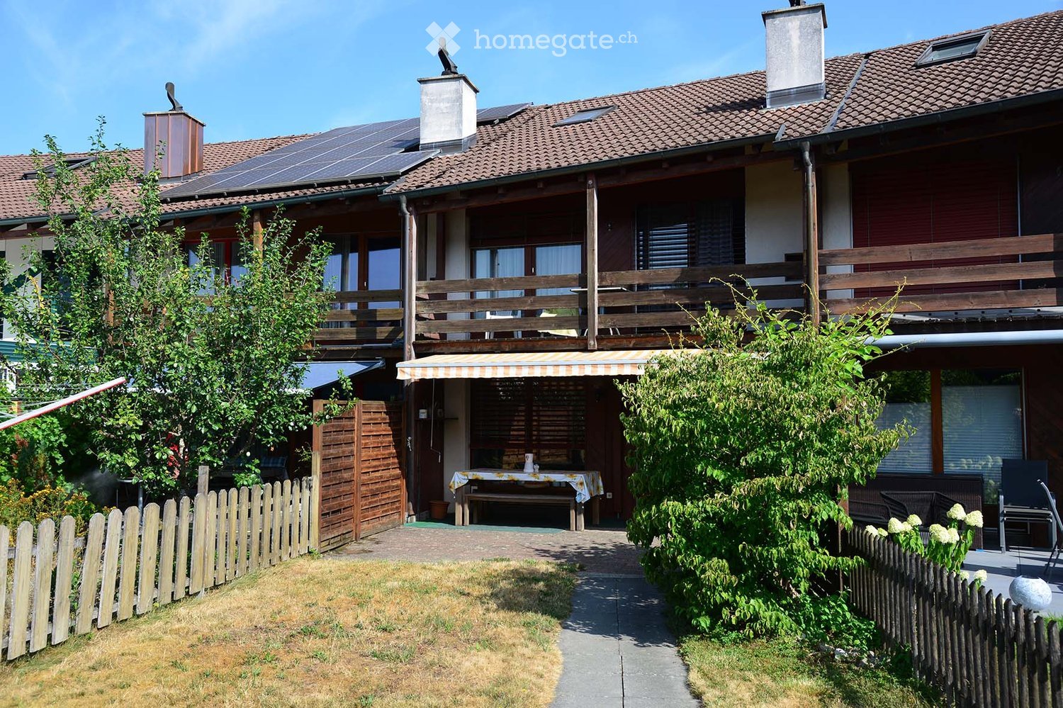 Two-story house with a tiled roof, solar panels, balconies, and a fenced garden area with outdoor furniture.