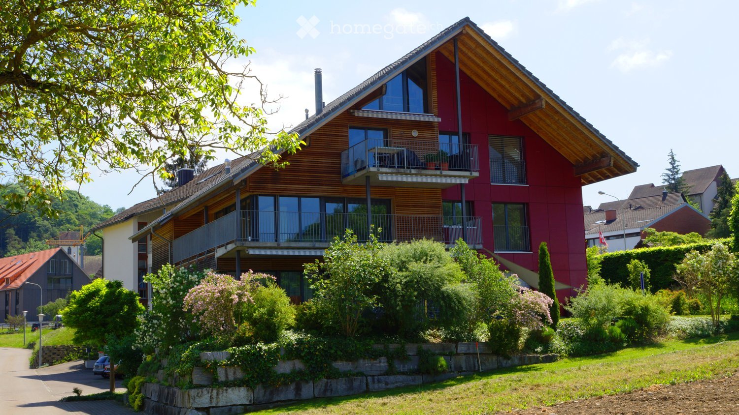 Modern two-story house with red and wooden exterior, multiple balconies, surrounded by greenery and trees.