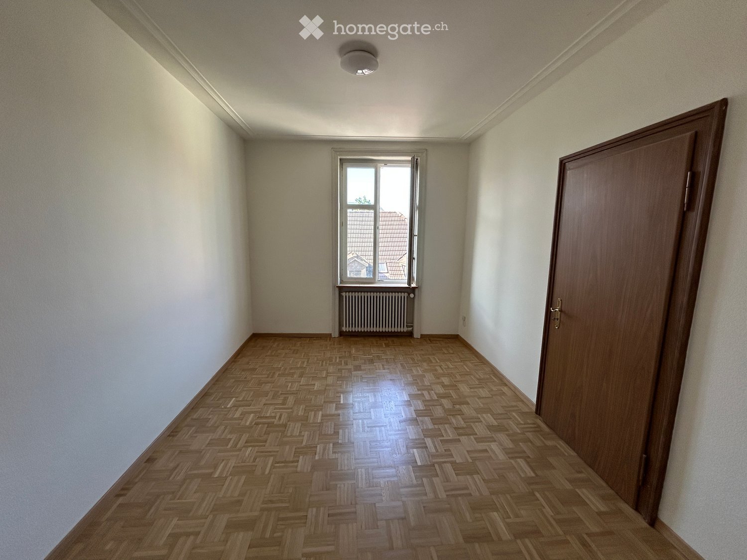 Empty room with wooden herringbone flooring, white walls, a single window with a radiator below, and a closed wooden door.