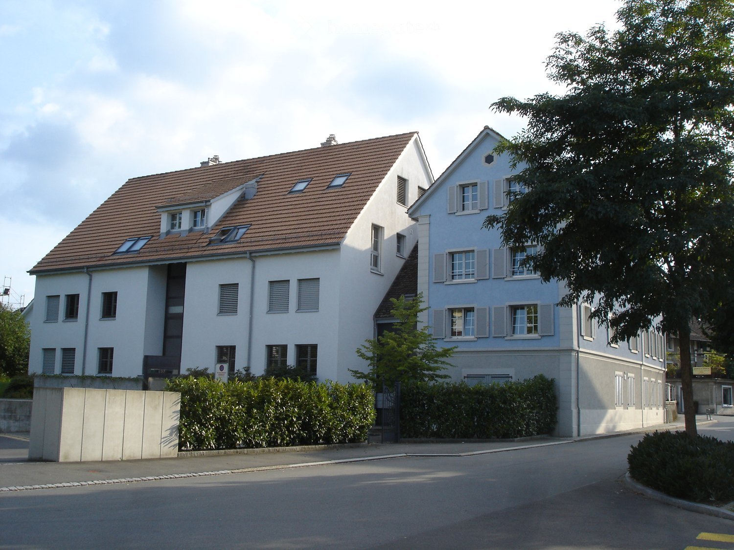 Two-story white house with brown tiled roof, multiple windows, and surrounding greenery.