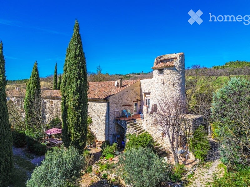 A rustic stone house with a tiled roof, surrounded by tall cypress trees and a well-maintained garden. The house features a balcony with outdoor seating and a small patio area.