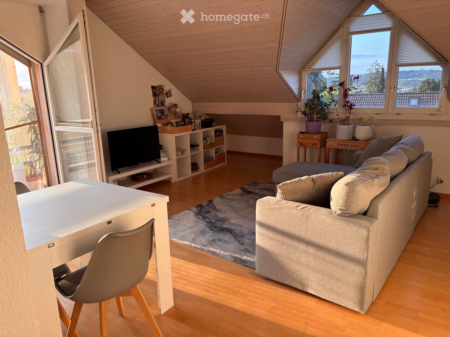 Bright living room with wooden floors, large windows offering a scenic view, a grey sofa, coffee table, modern chair, and a television on a shelf.