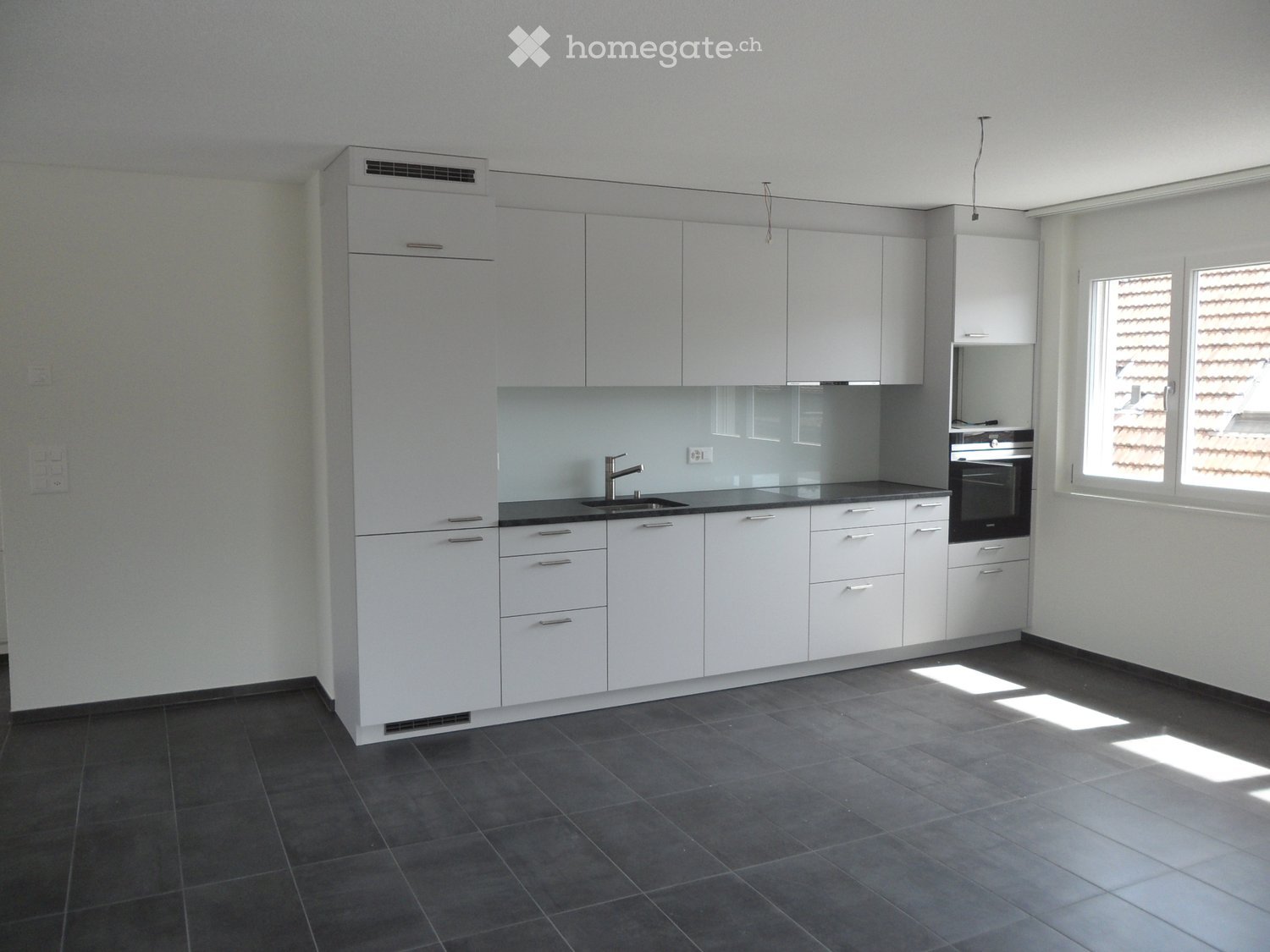 Modern kitchen with white cabinets, dark countertops, and a stainless steel sink. The space features built-in storage, a built-in oven, and large windows allowing natural light to enter.