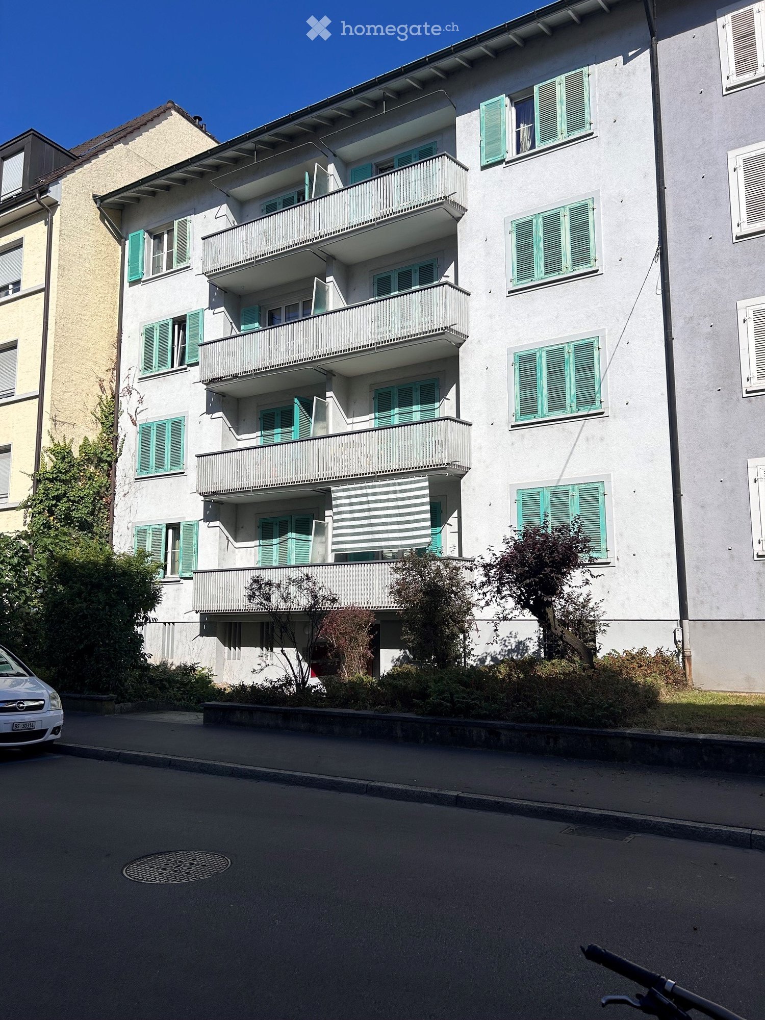 Multi-story residential building with green shutters, balconies, and a striped awning on the upper floor.