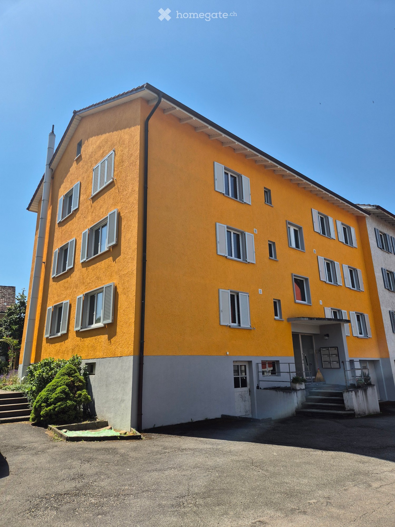 Two-story yellow house with white shutters and multiple windows, featuring a gray entrance area with steps and small plants.