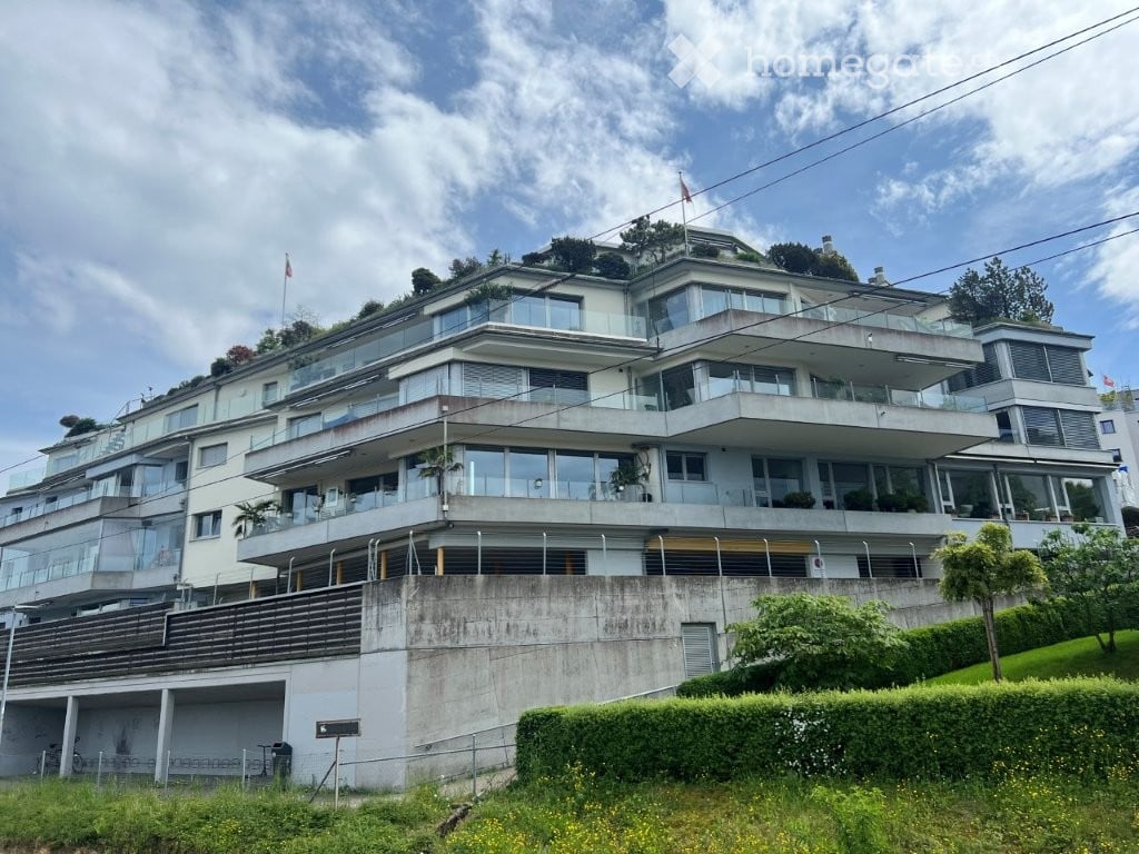 Modern multi-story building with large glass windows and balconies, situated on a raised ground floor with a landscaped garden in front.