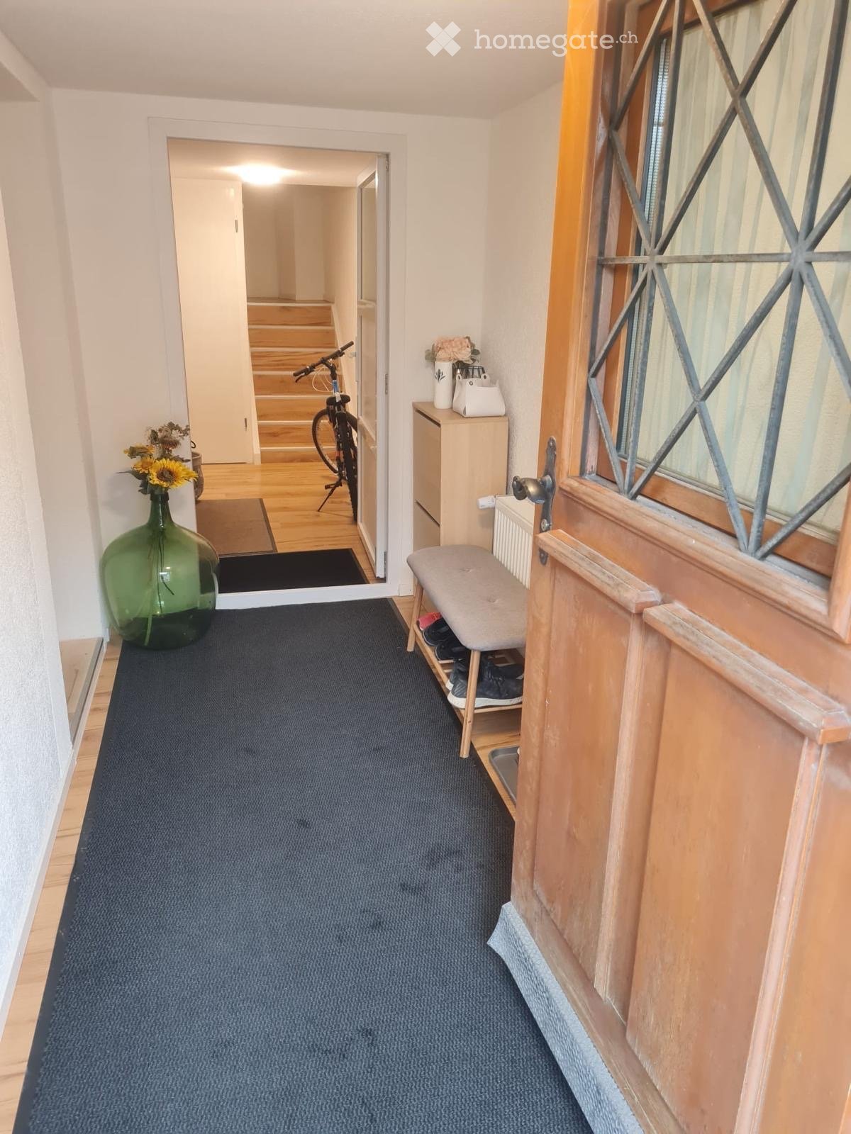 Hallway with blue carpet, wooden floor visible in adjacent area, small bench with shoes, bicycle, and decorative items including vases and flowers.