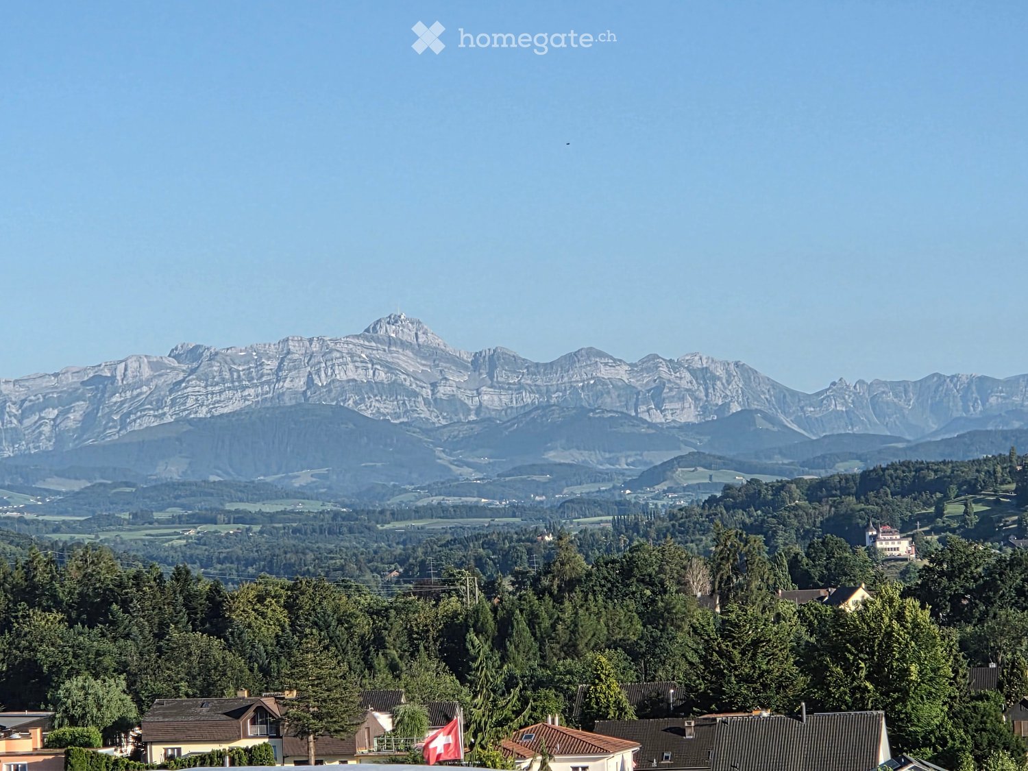 A panoramic view of a town nestled in a valley with a range of mountains in the background, featuring houses, trees, and a Swiss flag in the foreground.