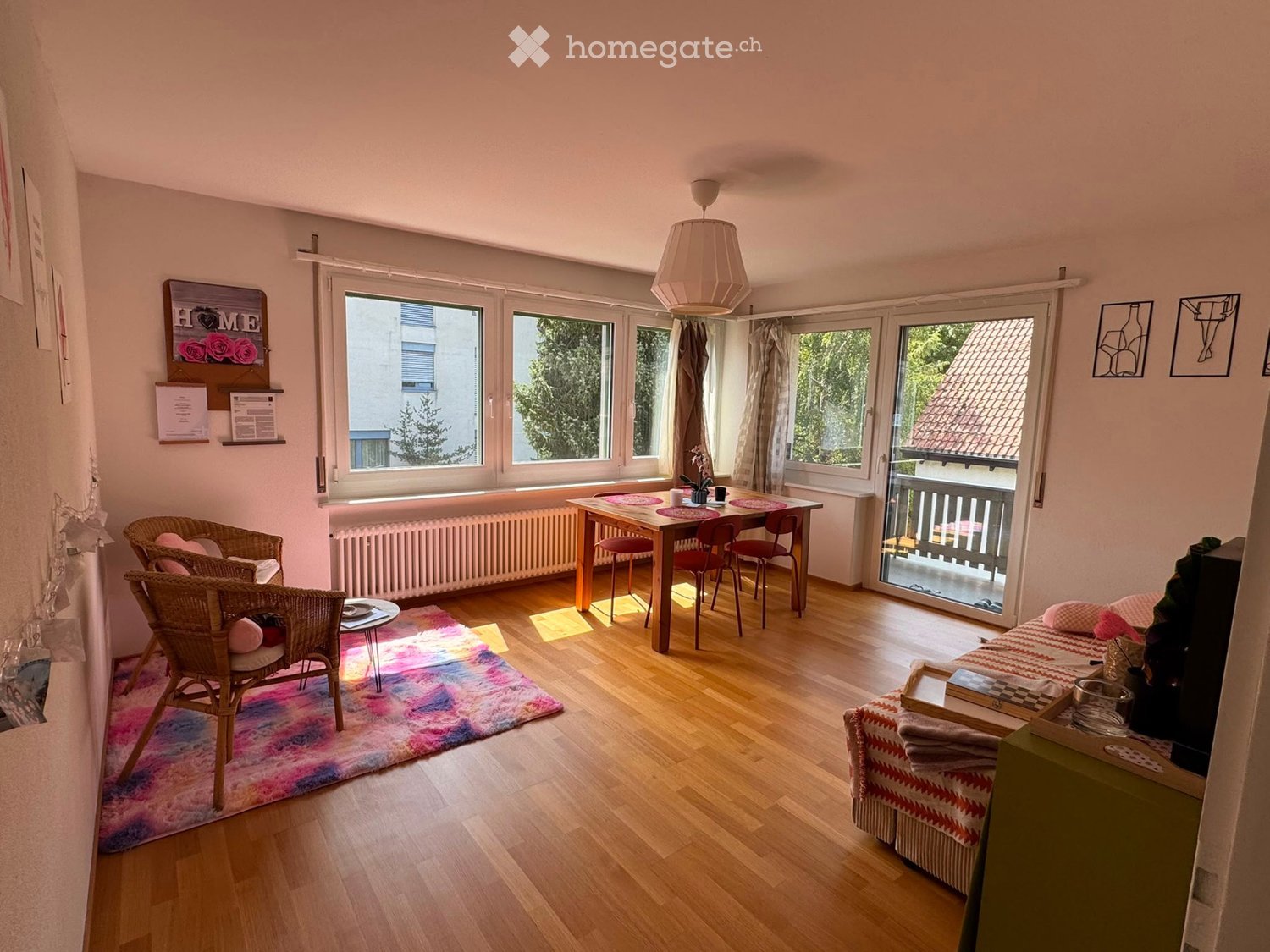 Bright living space with wooden flooring, large windows, a balcony, a sofa, a coffee table, a dining table with chairs, and wicker chairs.