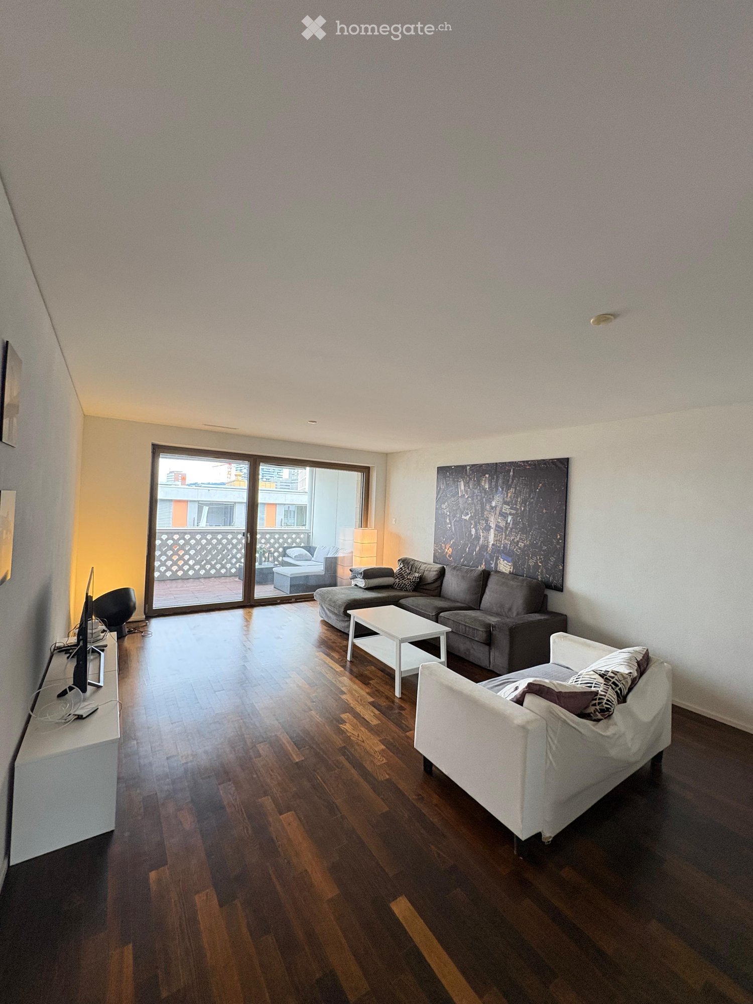 Modern living room with wooden flooring, large sliding glass doors leading to a balcony, a grey sectional sofa, a white coffee table, a white armchair, and a TV on a stand.