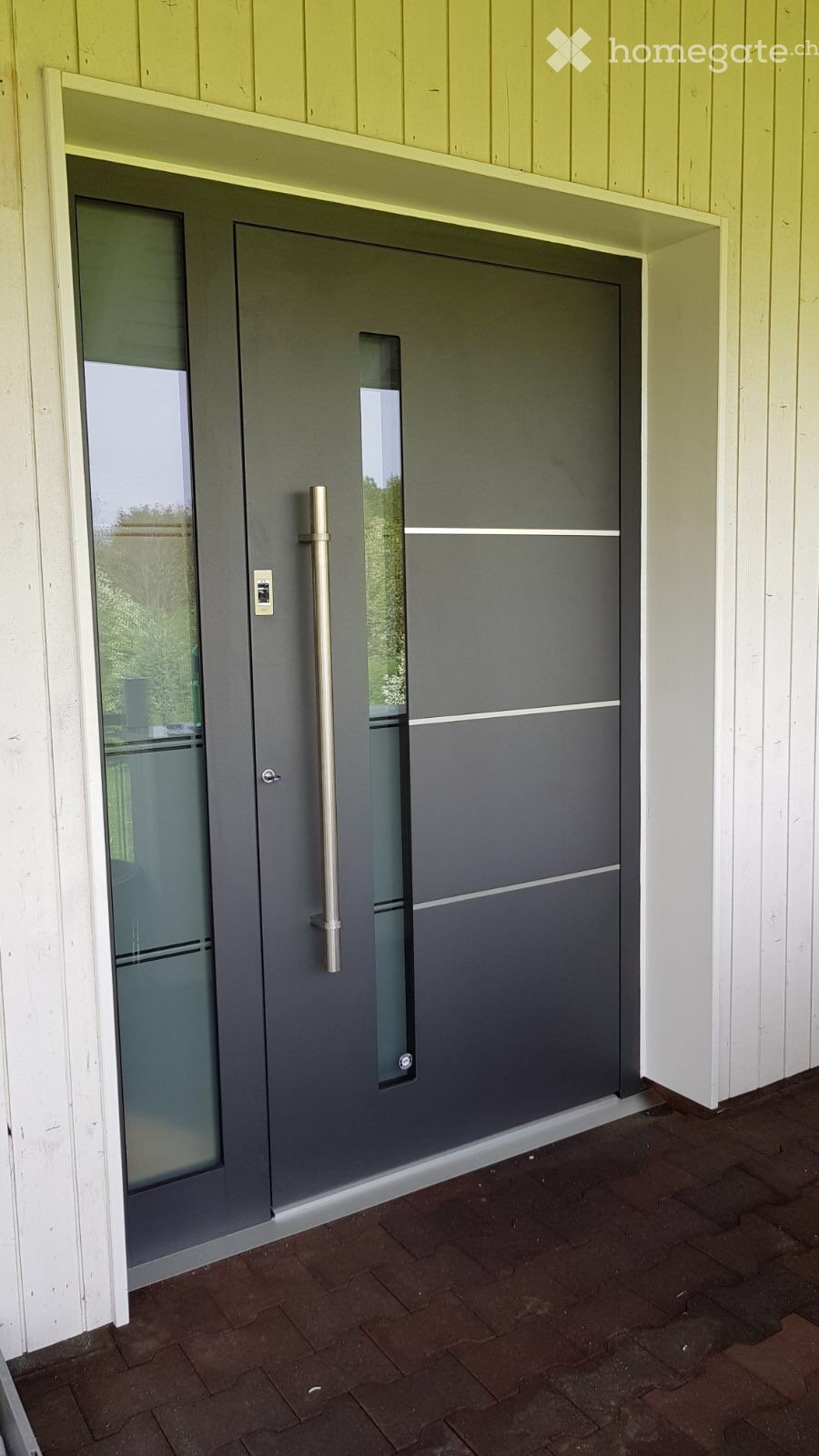 Modern grey front door with glass panels, set within a white wooden frame, featuring a brass handle and a small window panel above the door.