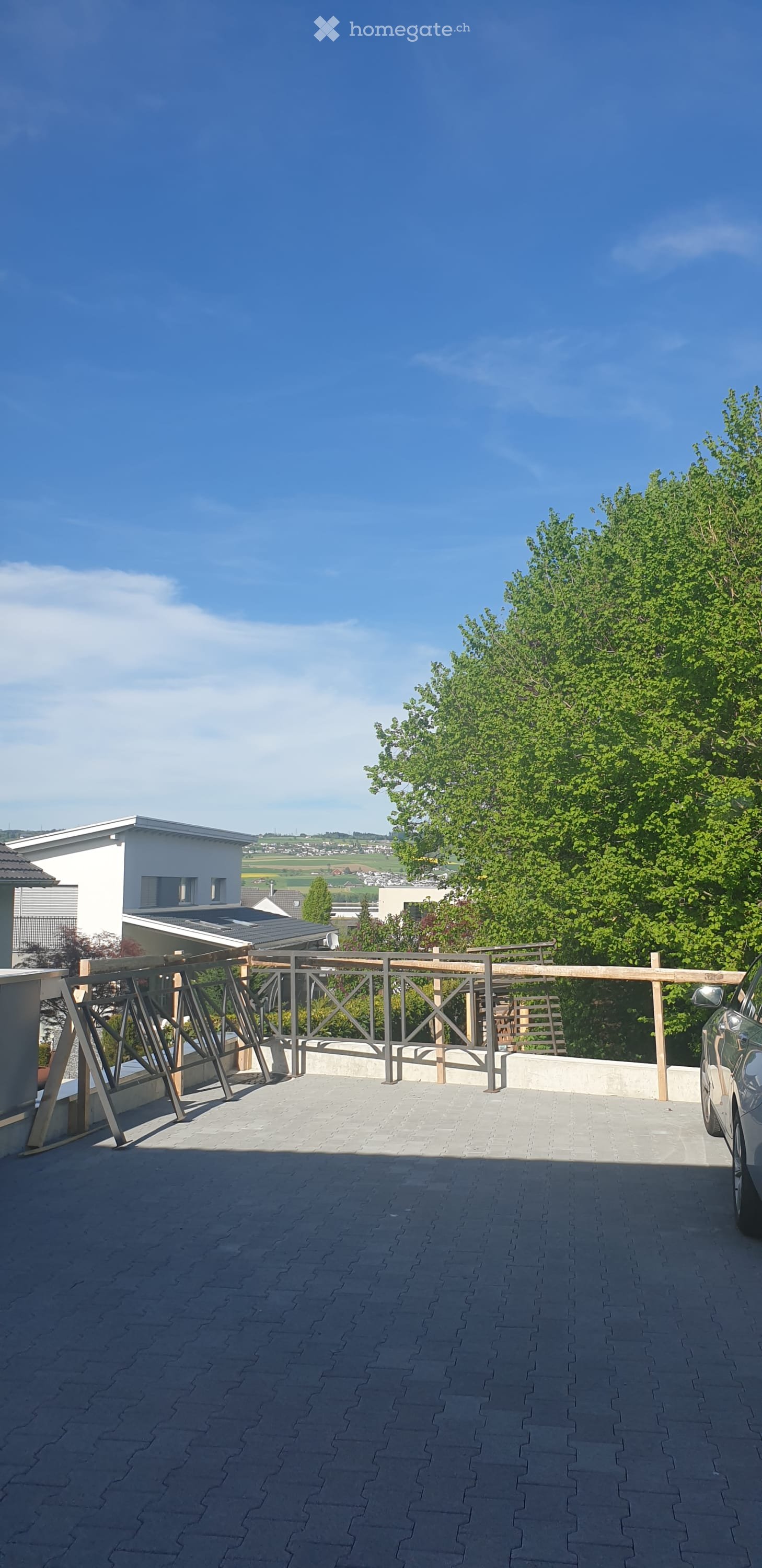 Balcony with wooden railing, paved floor, and a view of a house and greenery in the background.