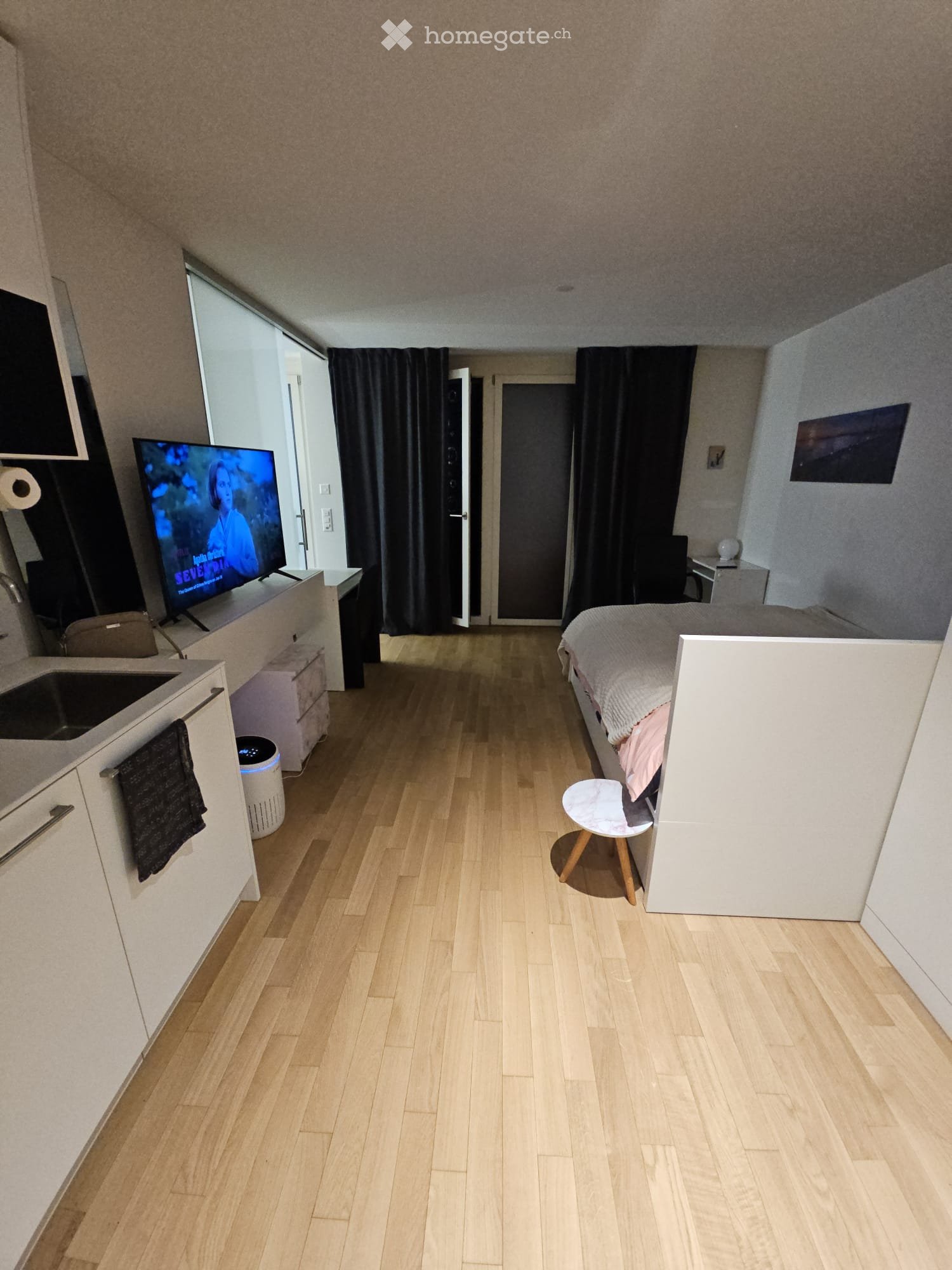 A compact studio apartment with a bed, a small sink area, a television, and a stool. The room features light wooden flooring and modern decor.