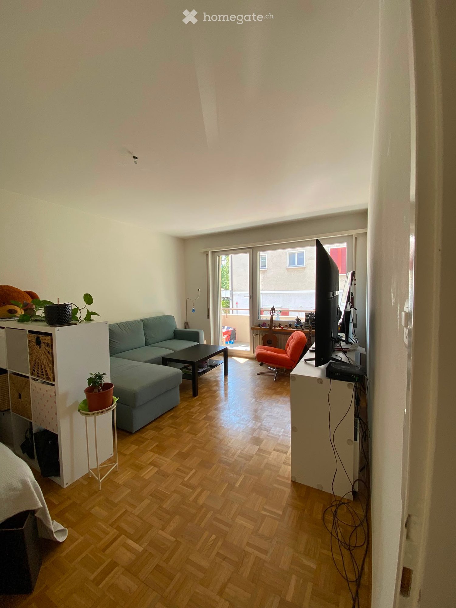 A modern living room with wooden flooring, a grey sectional sofa, a black coffee table, a red armchair, a guitar, a TV screen, and a small plant on a side table.