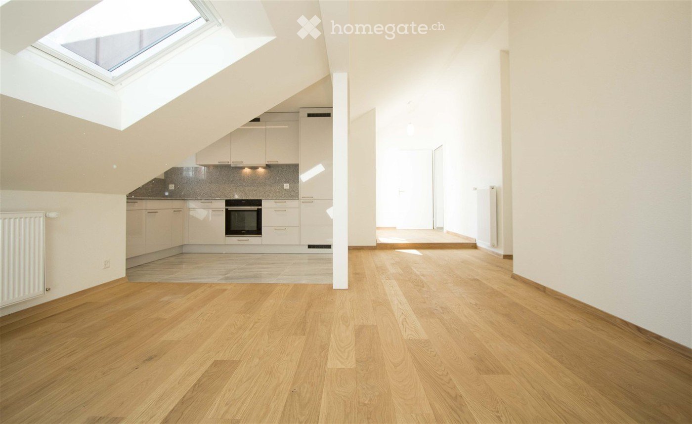 Bright, open loft-style room with wooden flooring, white walls, and a skylight. Adjacent kitchen area with white cabinets and stainless steel appliances.