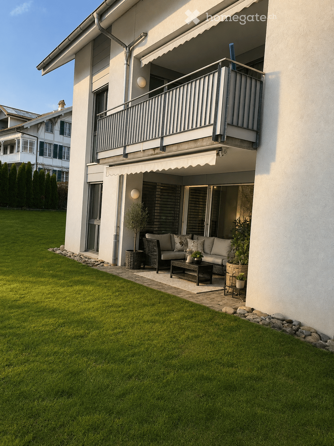 Well-maintained garden with a paved patio area featuring a sofa, coffee table, and potted plants. The patio is adjacent to a modern house with a balcony and awning.