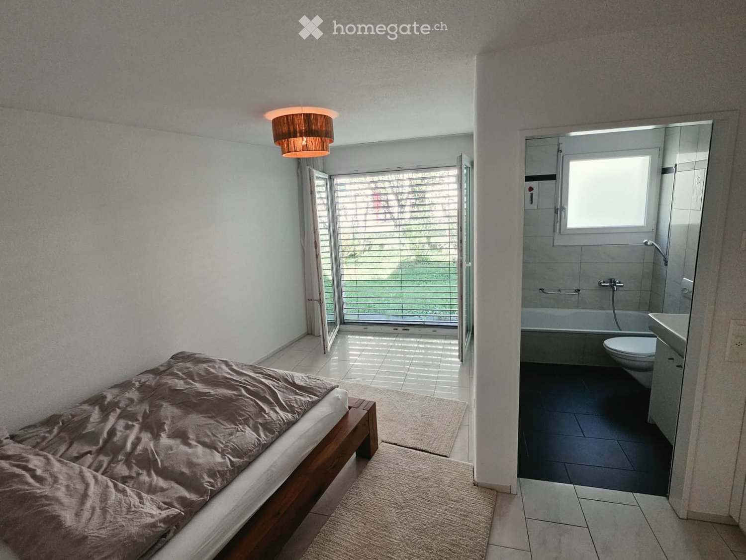 Modern bedroom with a wooden bed, beige bedding, and a connected bathroom featuring a toilet, sink, bathtub, and mirror. The room has large windows and a light fixture on the ceiling.