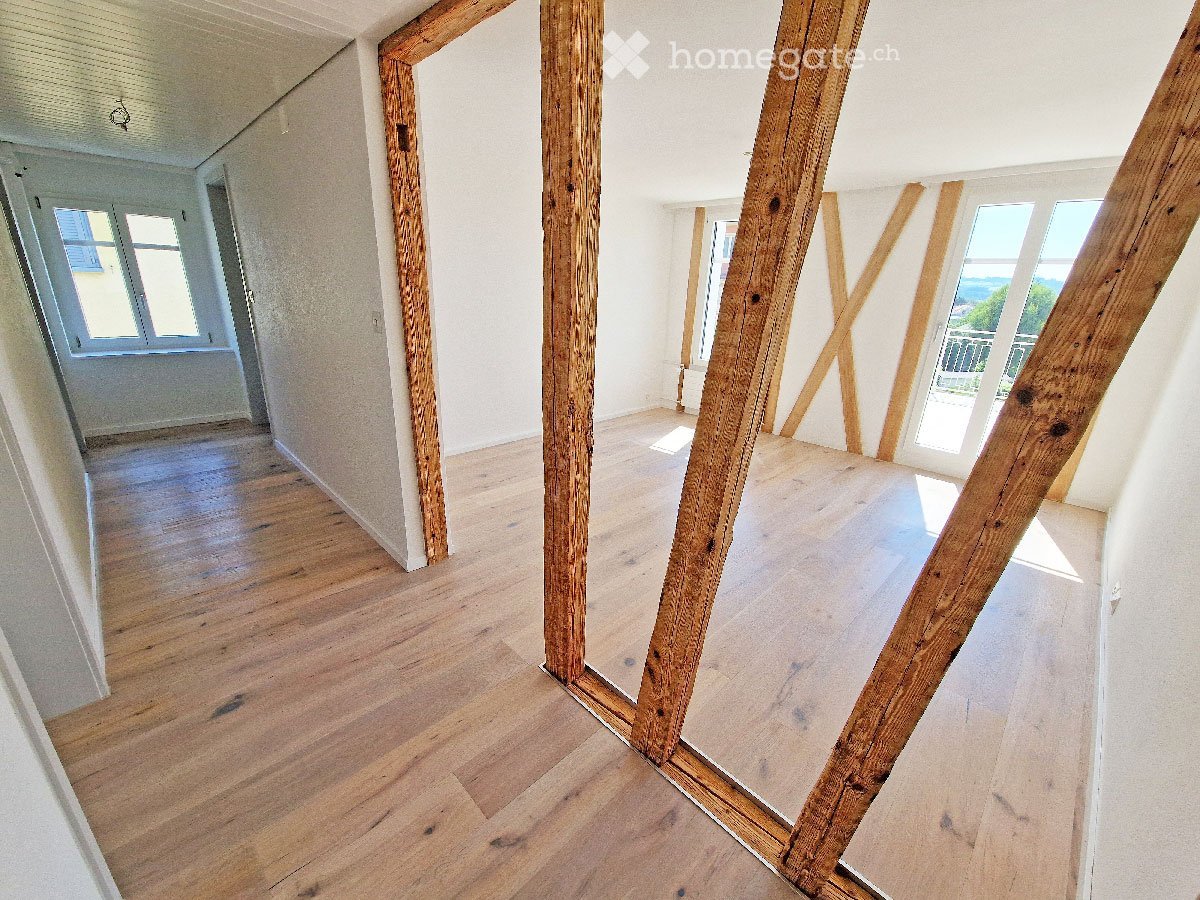 Empty room with wooden flooring and exposed wooden beams, large windows with views, and white walls.