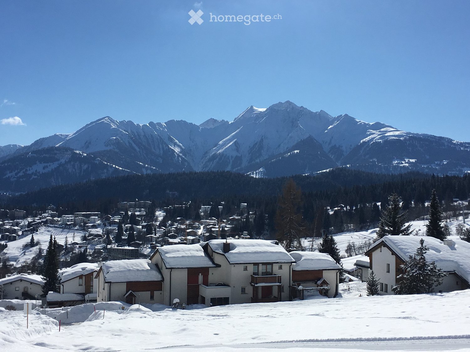 multiple snow covered chalets, surrounded by pine trees, mountains covered in snow in the background