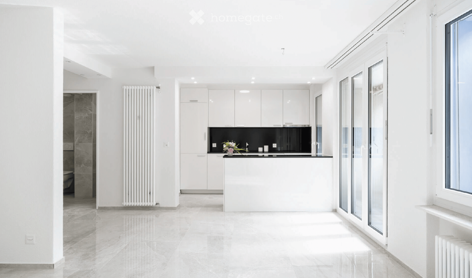 Modern, minimalist kitchen with white cabinets, a black countertop, and a sleek sink area. The space features large windows and a clean, open layout.
