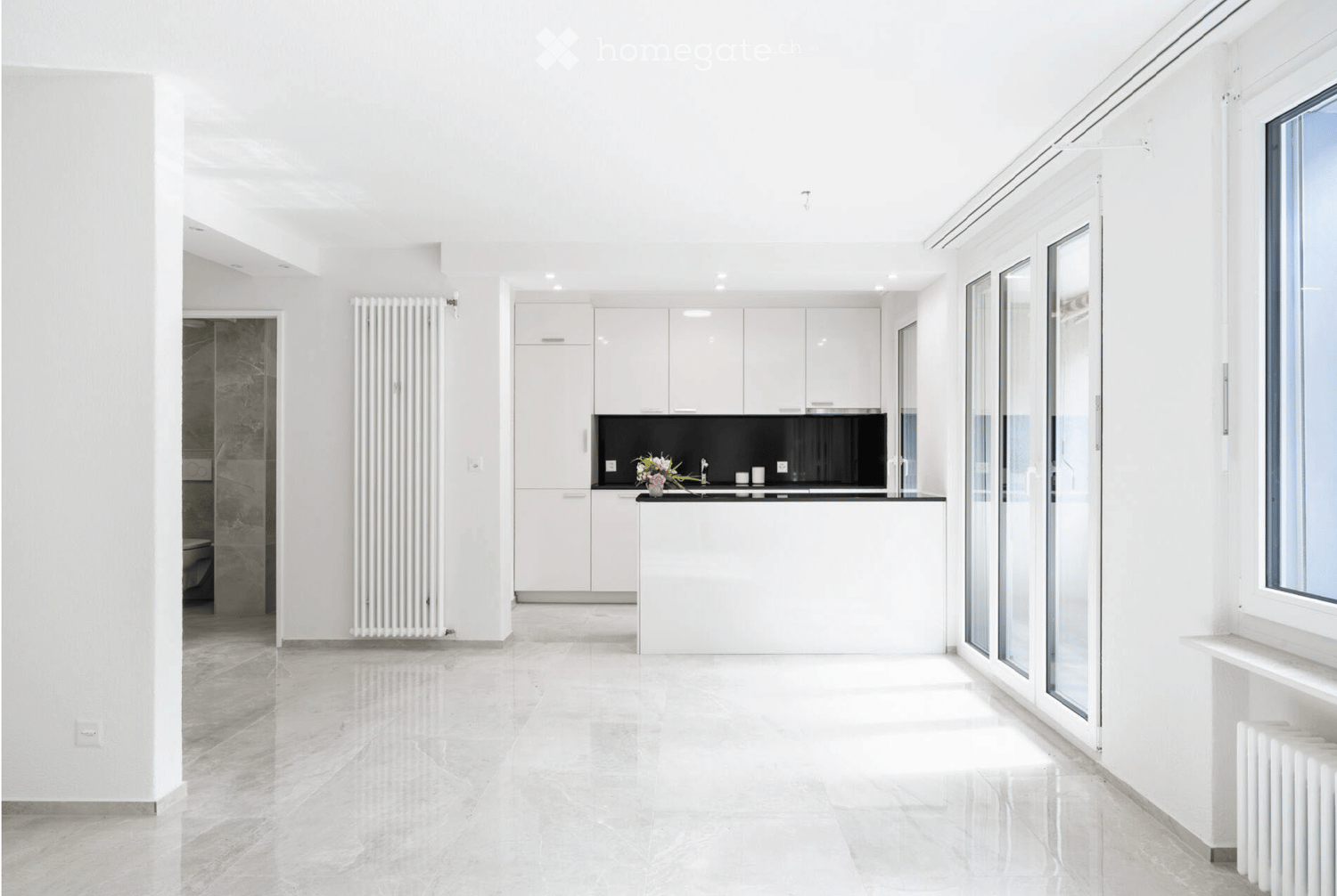 Modern, minimalist kitchen with white cabinets, a black countertop, and a sleek sink area. The space features large windows and a polished tile floor.
