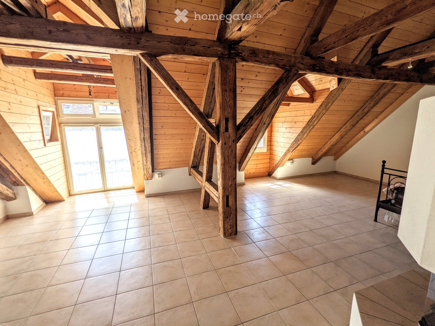 An empty attic space with wooden beams, tiled floor, and large windows, featuring exposed wooden beams and a sliding glass door leading outside.