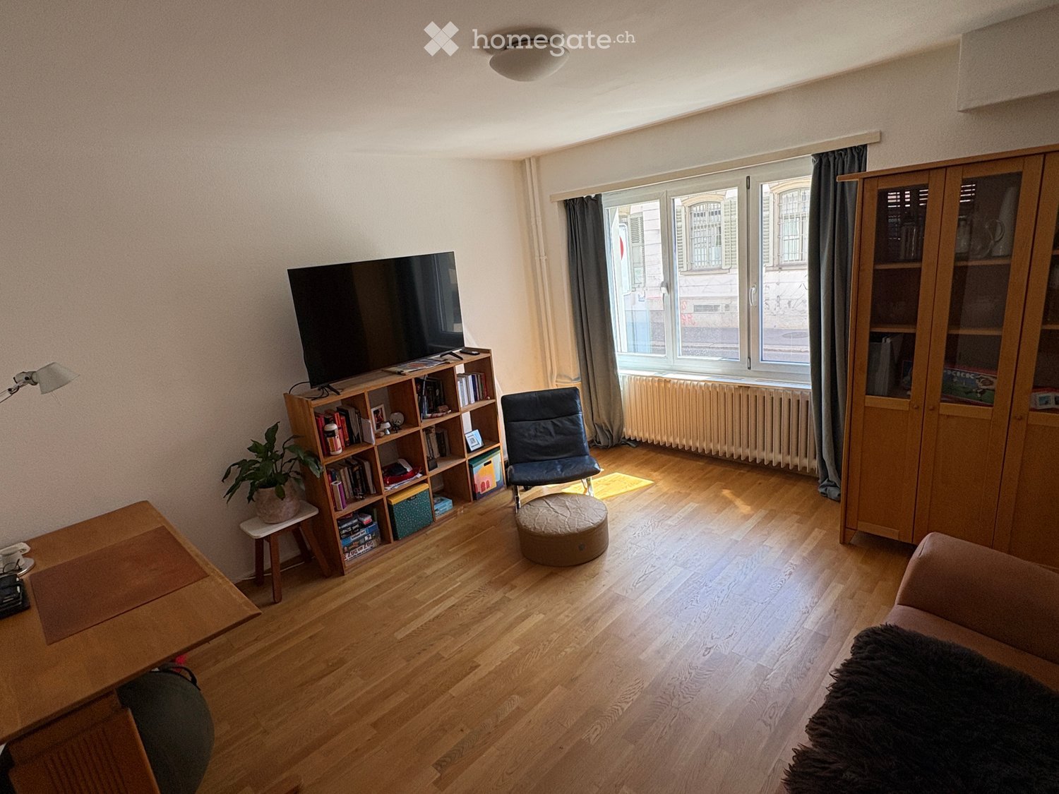 Living room with wooden flooring, large window with curtains, sofa, coffee table, bookshelf, chair, and television.