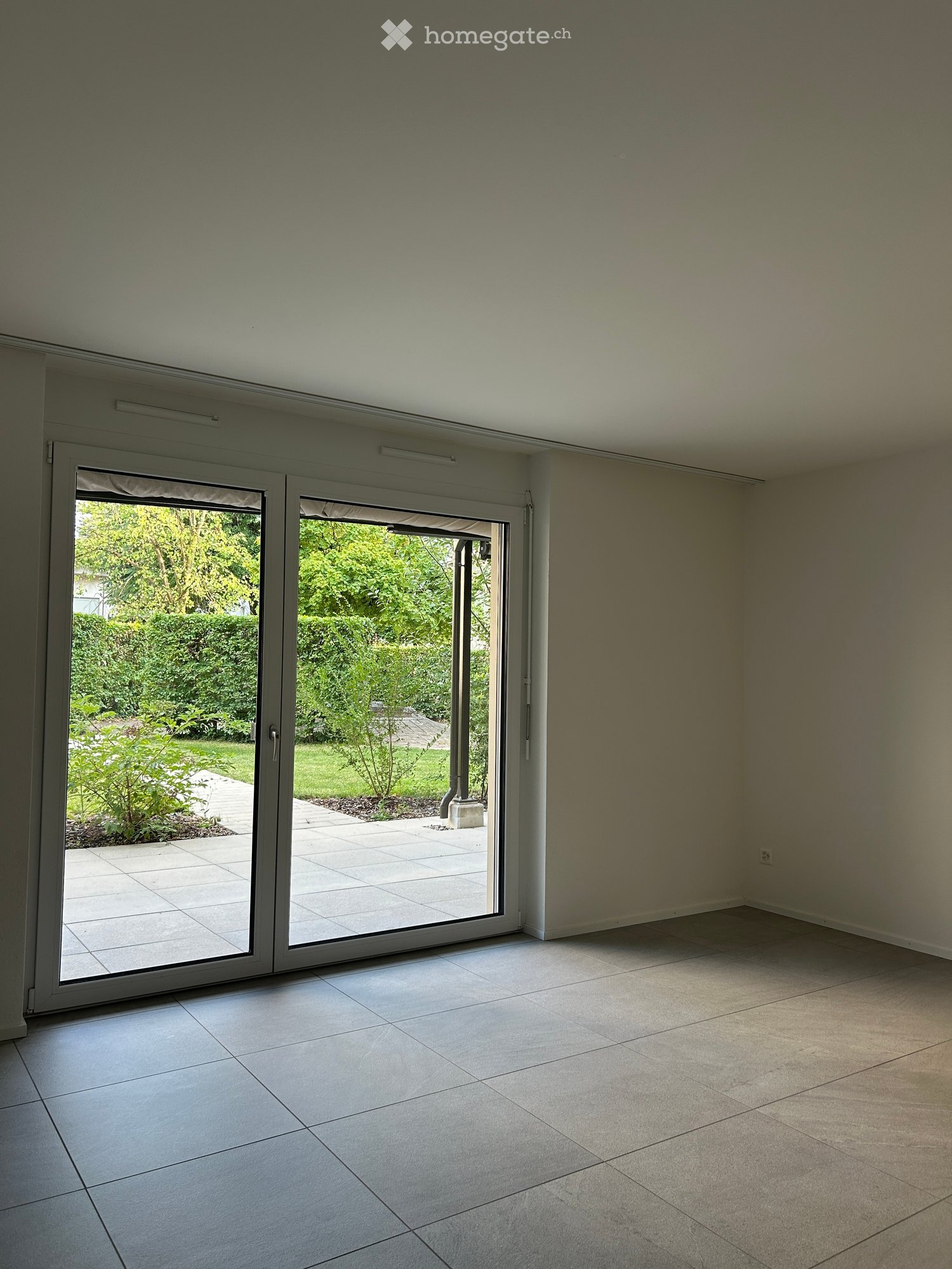 Empty room with large sliding glass doors leading to a garden, tiled floor, and neutral-colored walls.