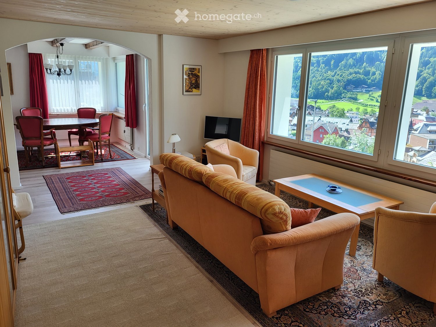 Bright living room with large windows offering a scenic view, furnished with a sofa, coffee table, armchair, dining table, chairs, television, and a table lamp.