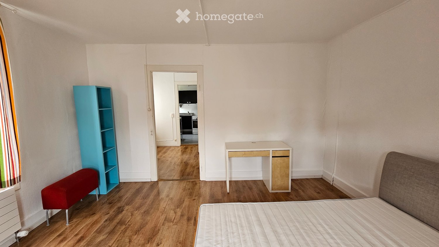 A furnished room with a bed, sofa, stool, shelf, table, and chair. The room has wooden flooring and white walls, with a view into an adjacent kitchen area.