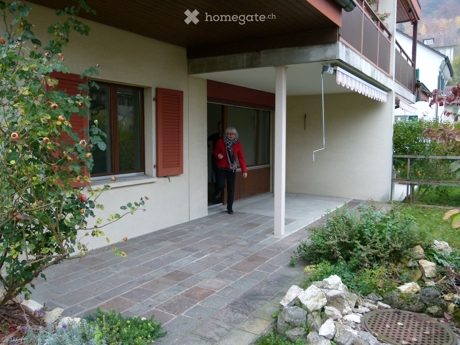 Garden area with tiled patio, small tree, shrubs, rocks, and a woman exiting the house.