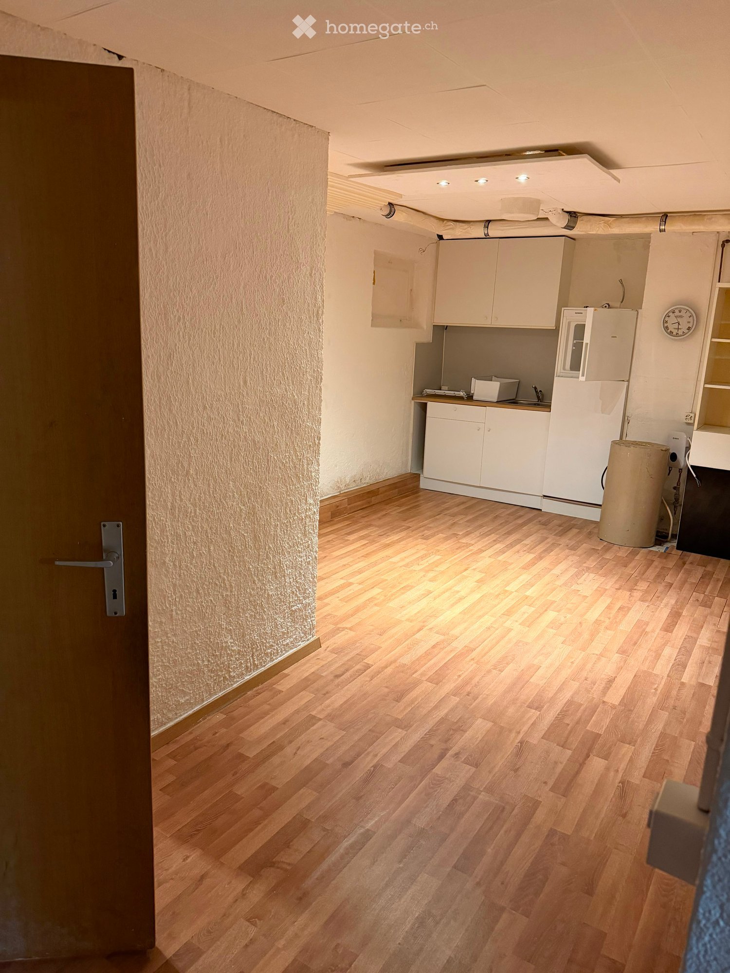 A small kitchen with white cabinets, a refrigerator, microwave, and sink, featuring wooden flooring and a door on the left side.