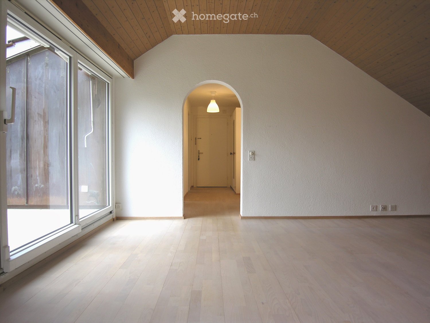 Empty room with wooden flooring, white walls, large windows, and an arched doorway leading to another room.
