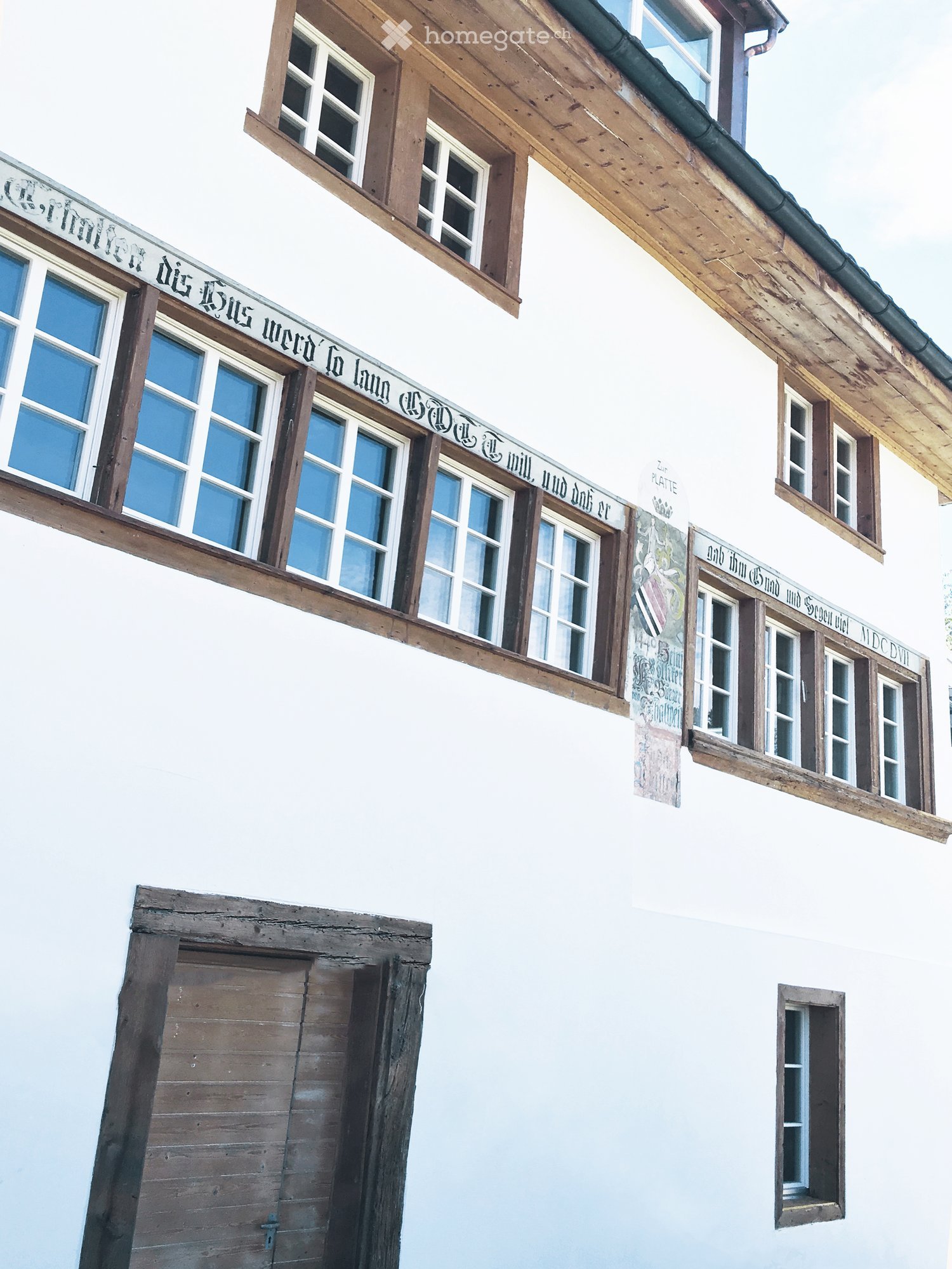 A two-story building with white walls, wooden beams, and multiple windows. The facade features historical inscriptions and a closed wooden door at the bottom left.