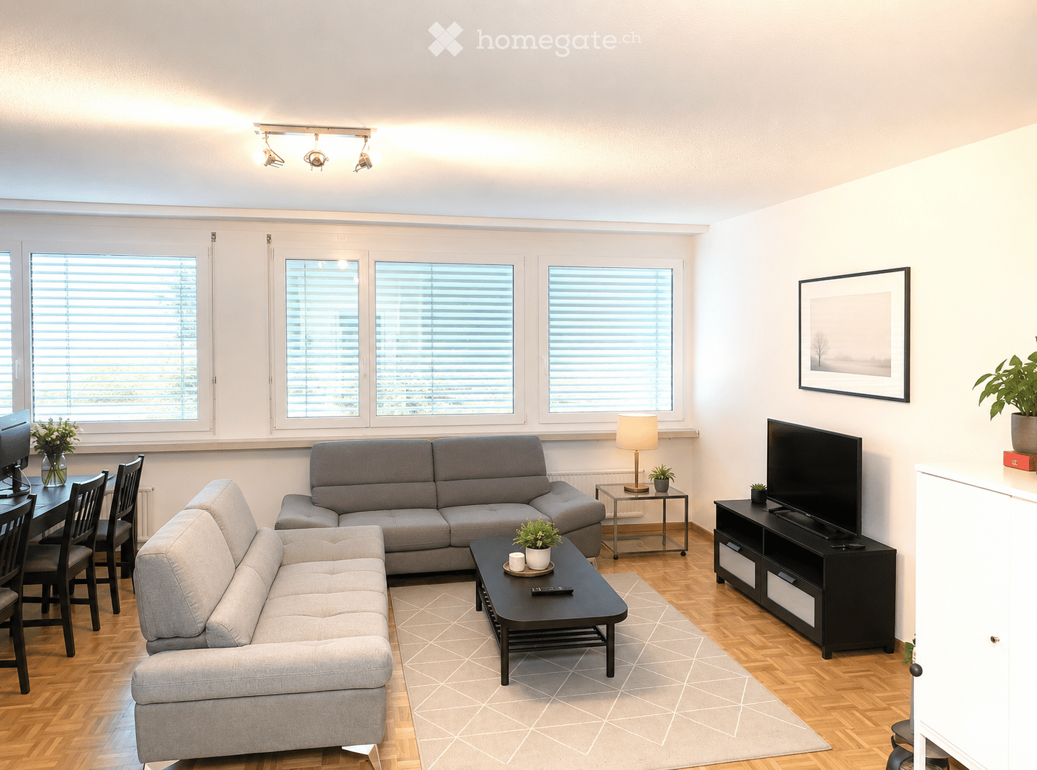 Modern living room with light-colored walls, wooden flooring, large windows with blinds, a sectional sofa, coffee table, dining table with chairs, TV stand, and decorative plants.