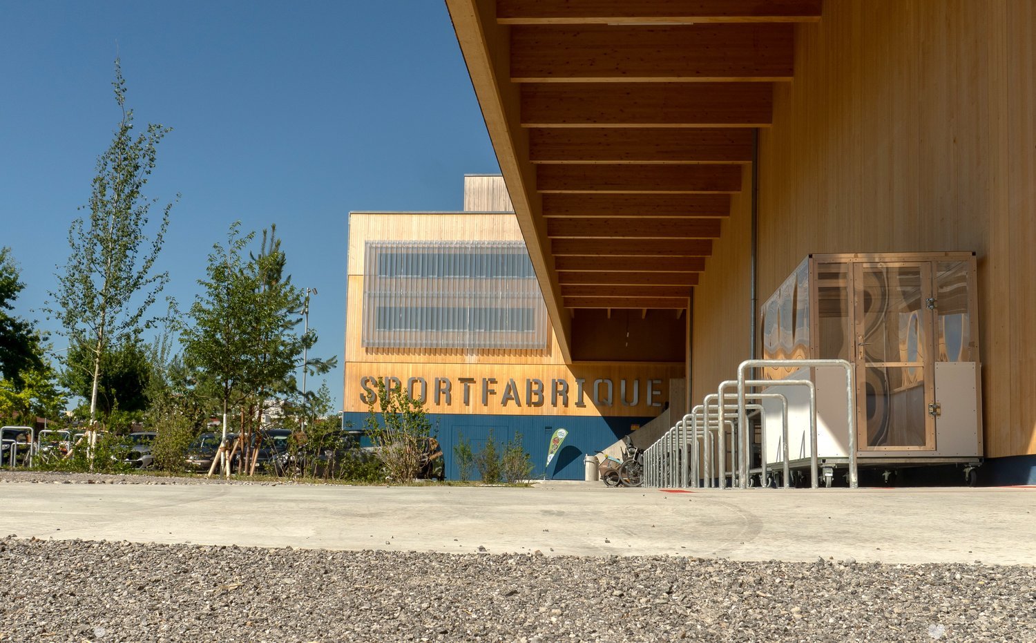 Modern building facade with wooden panels and large glass windows, labeled "SPORTFABRIQUE".